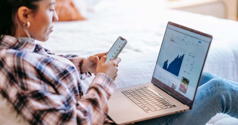 Dizüstü bilgisayar önünde telefonda grafik uygulaması kullanan kadın