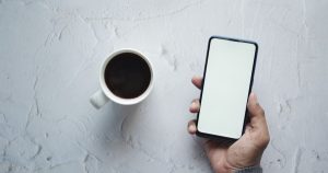 Beyaz zeminde kahve fincanı yanında boş ekranlı akıllı telefon tutan el