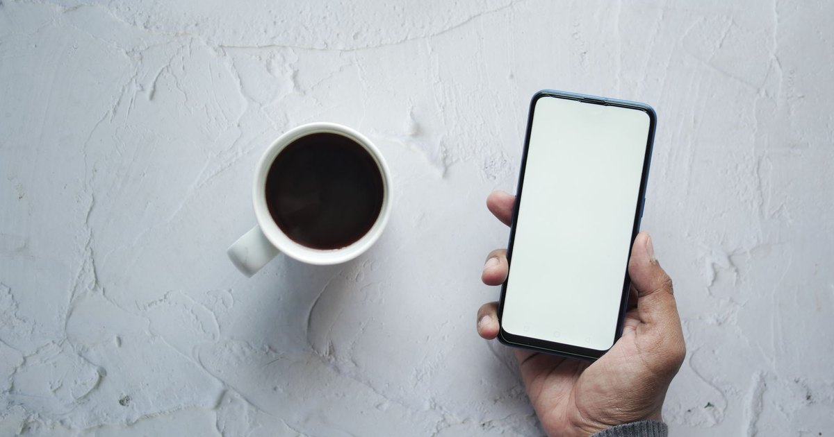Beyaz zeminde kahve fincanı yanında boş ekranlı akıllı telefon tutan el
