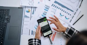 Dizüstü bilgisayar ve belgelerin yanında telefonda grafik inceleyen iş kadını