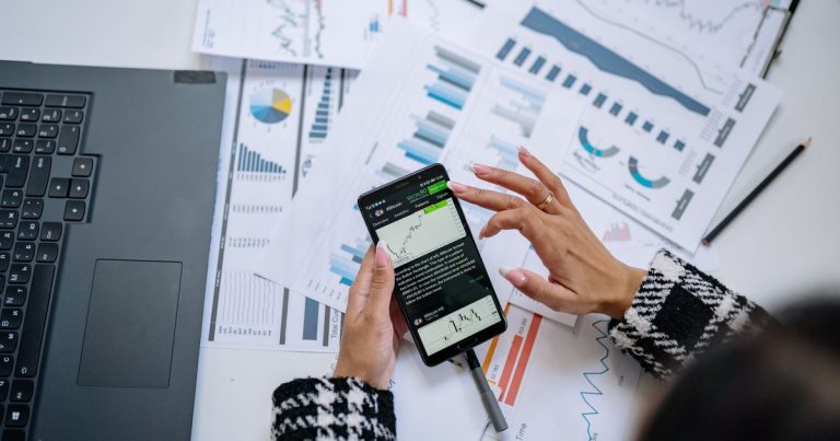 Dizüstü bilgisayar ve belgelerin yanında telefonda grafik inceleyen iş kadını